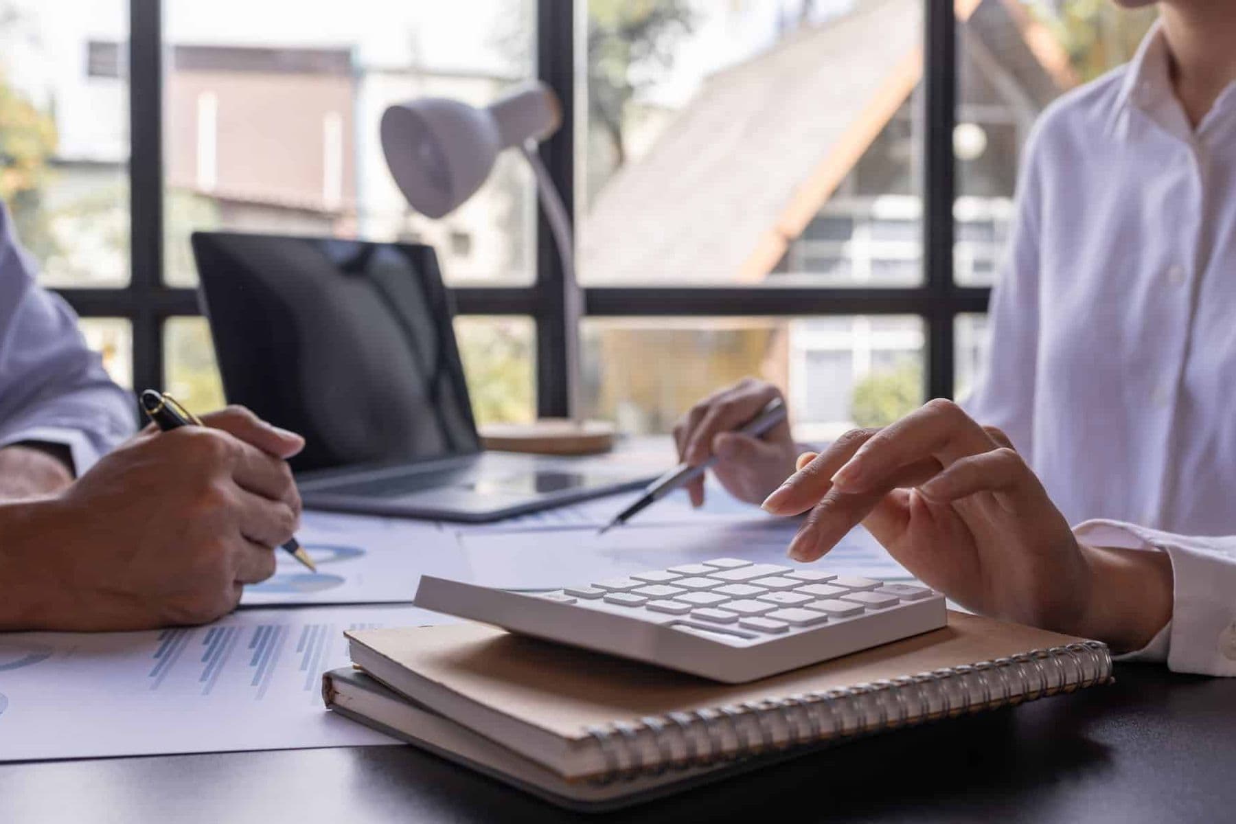 Two professionals reviewing financial documents with a calculator and laptop on a desk, in a bright office setting.