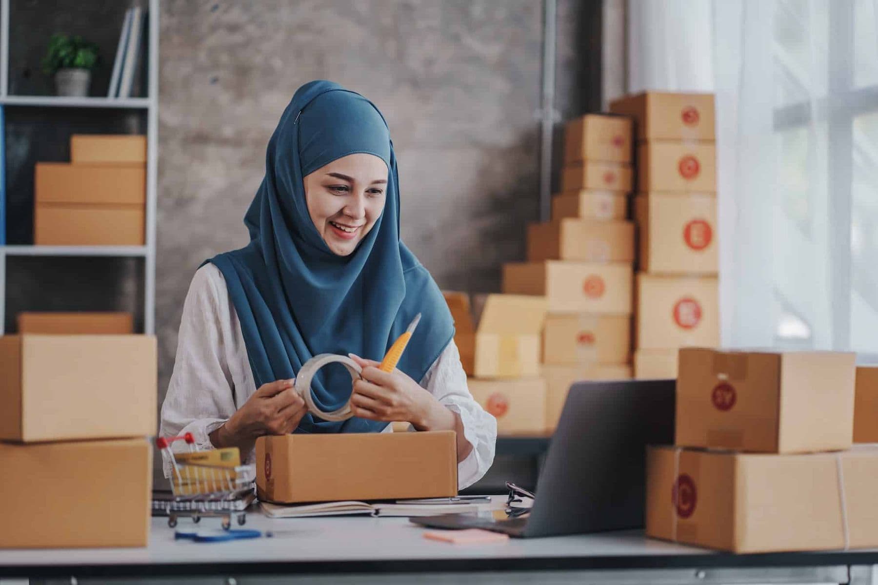 Smiling woman in hijab packing boxes for her small business, surrounded by shipping parcels and working on a laptop.