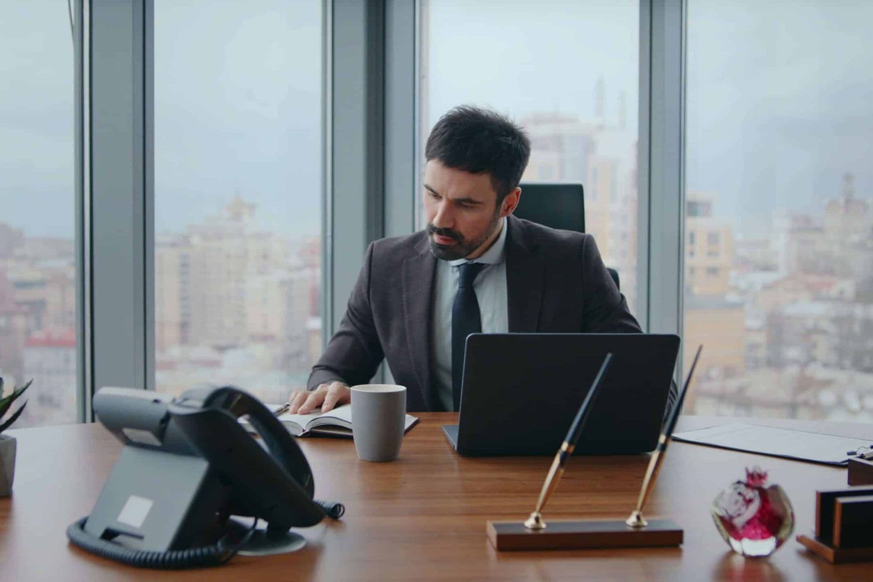 Business professional reviewing corporate compliance documents at a modern office desk with laptop and city view.